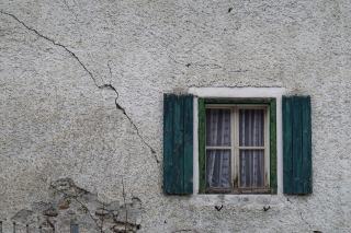 Façade enduite traversée par une longue fissure autour d'une fenêtre aux volets verts